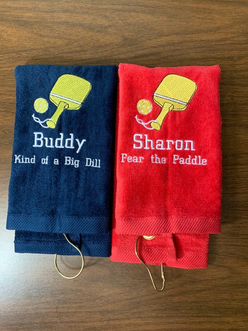 May include: Two personalised towels with embroidered paddleball rackets and text. The blue towel says "Buddy Kind of a Big Dill" and the red towel says "Sharon Fear the Paddle".