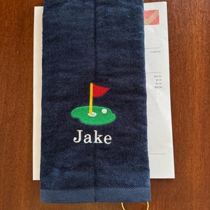 K&ouml;nnte beinhalten: Marineblaues Golfhandtuch mit goldenem Ring. Das Handtuch zeigt ein gesticktes Golfgr&uuml;n mit roter Fahne und dem Namen "Jake" in Wei&szlig;.
