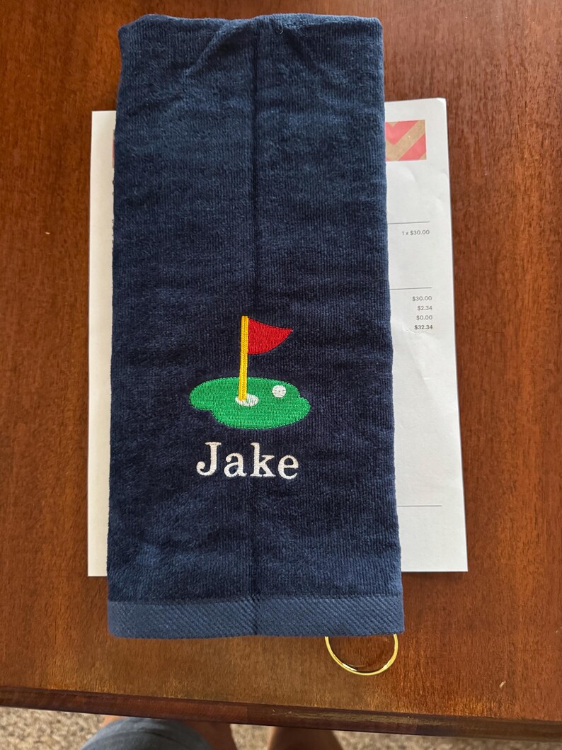 K&ouml;nnte beinhalten: Marineblaues Golfhandtuch mit goldenem Ring. Das Handtuch zeigt ein gesticktes Golfgr&uuml;n mit roter Fahne und dem Namen "Jake" in Wei&szlig;.