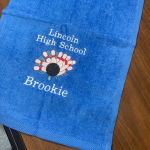 May include: Blue towel with white embroidery. The embroidery says "Lincoln High School" and features a bowling pin and ball graphic. The name "Brooke" is also embroidered on the towel.