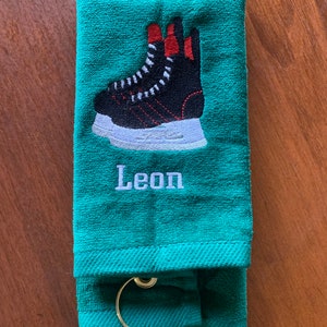May include: A green towel with a black and white hockey skate embroidered on it. The towel has a loop for hanging and the name "Leon" is embroidered below the skate.