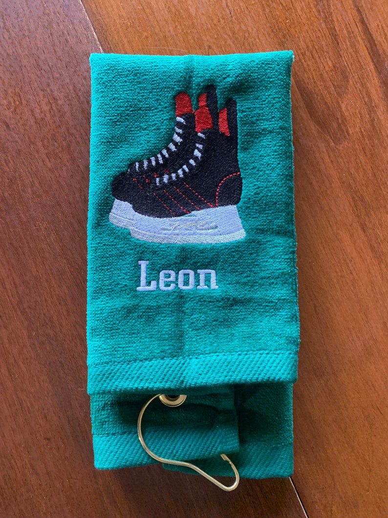 May include: A green towel with a black and white hockey skate embroidered on it. The towel has a loop for hanging and the name "Leon" is embroidered below the skate.