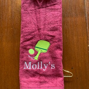 May include: A maroon towel with a white embroidered design of a green paddle and a yellow ball. The text "Molly's" is embroidered below the paddle and ball.