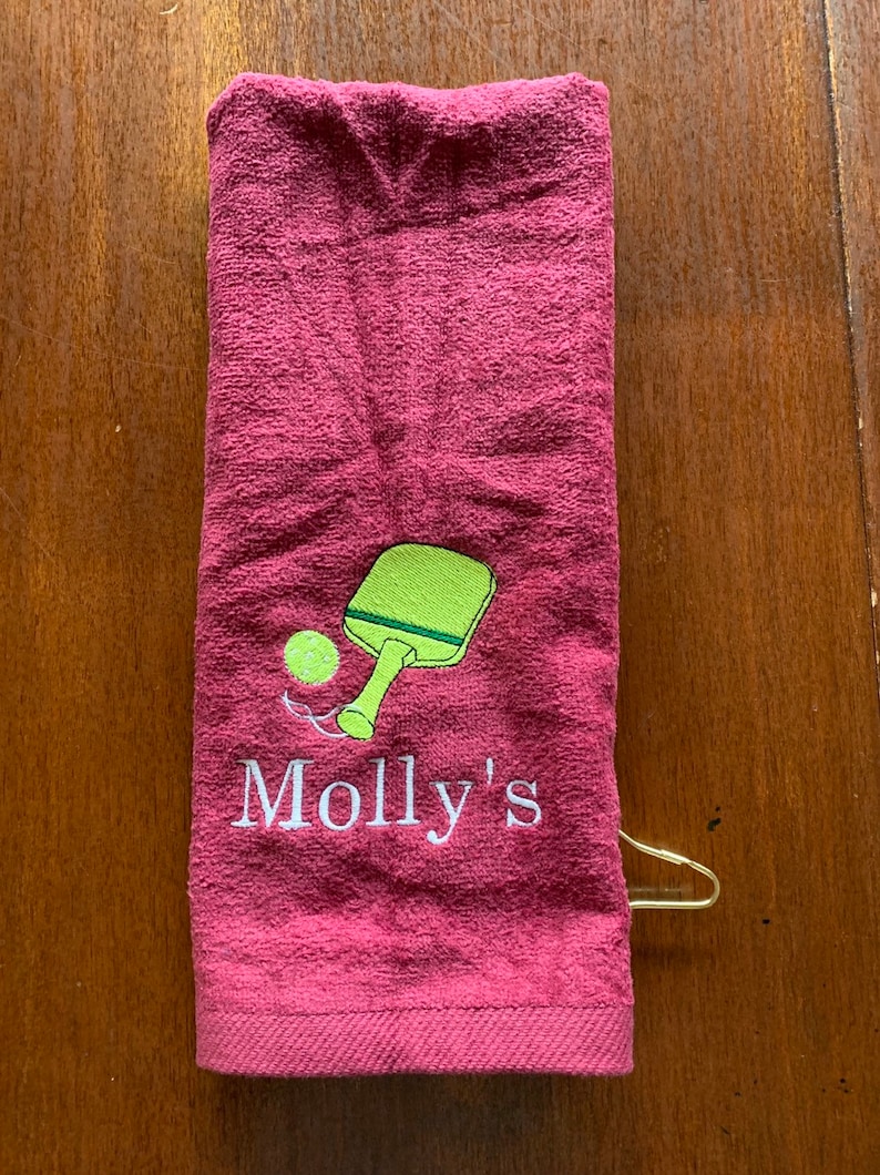 May include: A maroon towel with a white embroidered design of a green paddle and a yellow ball. The text "Molly's" is embroidered below the paddle and ball.