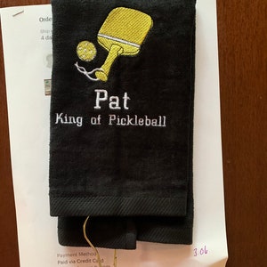 May include: Black towel with embroidered yellow pickleball paddle and ball, and the text "Pat King of Pickleball".