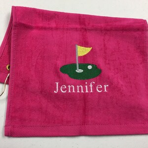 K&ouml;nnte beinhalten: Ein pinkfarbenes Golfhandtuch mit goldenem Ring und einem Golfplatz-Design, das in Gr&uuml;n, Gelb und Wei&szlig; gestickt ist. Der Name "Jennifer" ist darunter in Wei&szlig; gestickt.