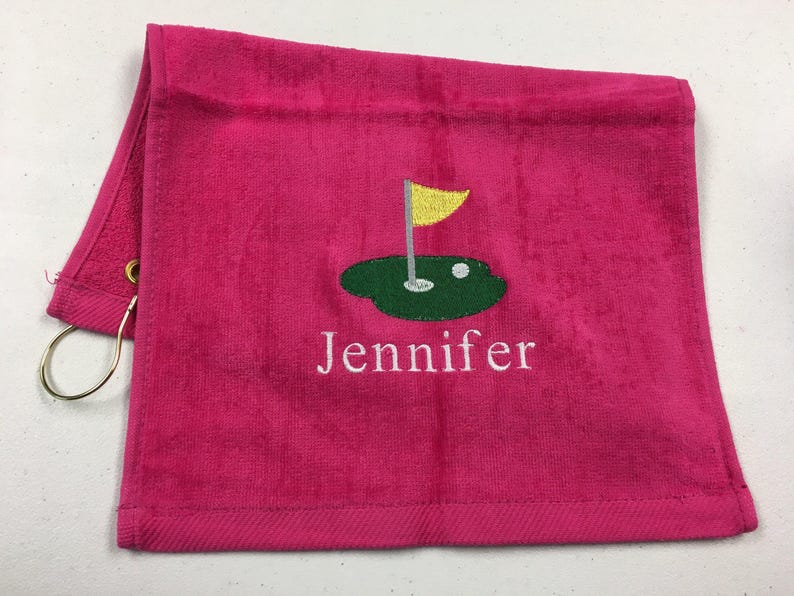 K&ouml;nnte beinhalten: Ein pinkfarbenes Golfhandtuch mit goldenem Ring und einem Golfplatz-Design, das in Gr&uuml;n, Gelb und Wei&szlig; gestickt ist. Der Name "Jennifer" ist darunter in Wei&szlig; gestickt.