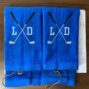 K&ouml;nnte beinhalten: Zwei blaue Golfhandt&uuml;cher mit den gestickten Initialen "L D" und gekreuzten Golfschl&auml;gern. Jedes Handtuch hat einen Metallring zum Aufh&auml;ngen. Die Handt&uuml;cher sind gefaltet und auf einer braunen Oberfl&auml;che gestapelt.