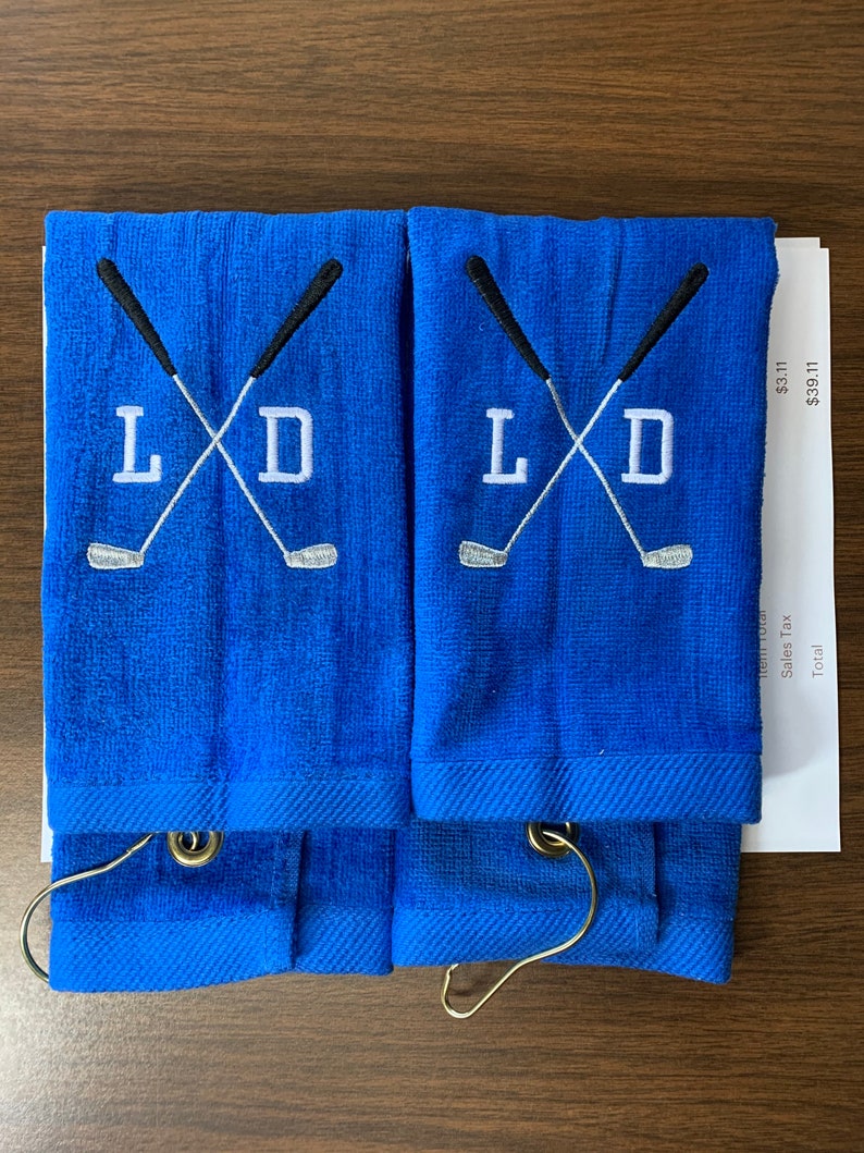 K&ouml;nnte beinhalten: Zwei blaue Golfhandt&uuml;cher mit den gestickten Initialen "L D" und gekreuzten Golfschl&auml;gern. Jedes Handtuch hat einen Metallring zum Aufh&auml;ngen. Die Handt&uuml;cher sind gefaltet und auf einer braunen Oberfl&auml;che gestapelt.