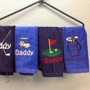 K&ouml;nnte beinhalten: Vier Golf-Handt&uuml;cher h&auml;ngen an einem Gestell. Die Handt&uuml;cher sind marineblau und blau und mit Golfmotiven und den W&ouml;rtern "Daddy" und "Grandpa" bestickt. Die Designs umfassen gekreuzte Golfschl&auml;ger, einen Golfwagen, ein Golfloch und eine Golftasche.