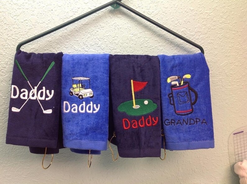 K&ouml;nnte beinhalten: Vier Golf-Handt&uuml;cher h&auml;ngen an einem Gestell. Die Handt&uuml;cher sind marineblau und blau und mit Golfmotiven und den W&ouml;rtern "Daddy" und "Grandpa" bestickt. Die Designs umfassen gekreuzte Golfschl&auml;ger, einen Golfwagen, ein Golfloch und eine Golftasche.