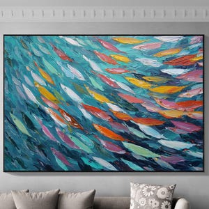 以下が含まれることがあります： 大きな額入りの抽象画で、魚の群れを描いています。この作品は、深い青色の背景に、青、オレンジ、黄色、ピンク、緑の色合いの鮮やかな魚の配列が特徴です。絵は灰色のソファの上に飾られています。