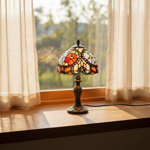 Puede incluir: Una lámpara de mesa de vidrieras con un diseño floral colorido. La lámpara tiene una base de color bronce y una pantalla con piezas de vidrio rojas, naranjas, verdes y blancas. La lámpara está colocada sobre una superficie de madera cerca de una ventana con cortinas transparentes.