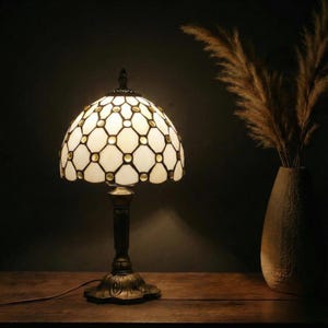 May include: A decorative table lamp with a stained glass shade featuring a white and gold geometric pattern. The lamp has a dark bronze base and stem, and is placed on a wooden surface. A vase with dried plants is in the background.