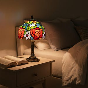 May include: A stained glass table lamp with a floral design. The lamp features a colorful shade with red, orange, white, green, and blue glass pieces. The base is dark brown and ornate. An open book sits on the wooden nightstand next to the lamp.