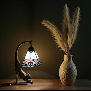 Puede incluir: Lámpara decorativa estilo Tiffany con base en forma de gato. La lámpara tiene una pantalla de vidrieras con detalles azules, blancos y rojos. Un jarrón con hierba de la pampa seca está a la derecha. La lámpara y el jarrón están sobre una superficie de madera.