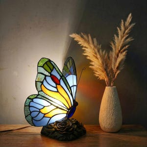May include: A stained glass butterfly lamp with a colorful design, featuring yellow, blue, green, and red hues. The lamp sits on a dark base with a rose design. A vase with dried plants is next to the lamp.