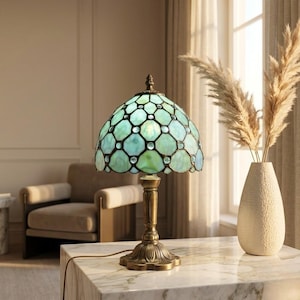 May include: A Tiffany-style table lamp with a green and blue stained glass shade. The lamp has a bronze-coloured base and a decorative finial. The lamp is on a marble-topped table, with a vase of dried flowers in the background.