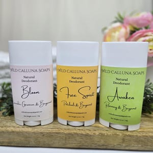 May include: Three natural deodorant sticks with white tops and colorful labels. The labels read "Bloom," "Free Spirit," and "Awaken," with ingredient lists below. The sticks are displayed on a wooden surface.