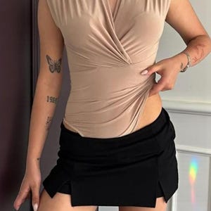 May include: A beige sleeveless bodysuit with a V-neckline and a draped design. Paired with a black mini skirt featuring a slit on the side. The outfit is simple and stylish, suitable for casual wear.
