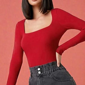 May include: A long-sleeved, red bodysuit with a square neckline. The top is paired with high-waisted, dark gray denim jeans. The jeans have a paper bag waist and two white buttons. The background is a solid coral color.