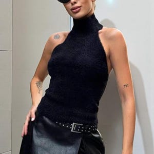 Op de afbeelding: Zwarte haltertop met een hoge halslijn en een fuzzy textuur. Gedragen met een zwarte leren wikkelrok met een riem met studs. De persoon draagt een zwarte zonnebril.