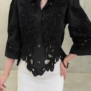 Può includere: Camicia nera con design floreale traforato. La camicia ha maniche a tre quarti e un colletto. Il fondo della camicia ha un bordo smerlato. Abbinata a una minigonna bianca.