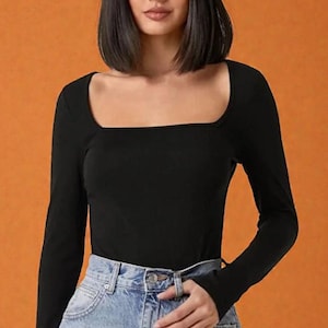 Könnte beinhalten: Schwarzes Langarm-Top mit quadratischem Ausschnitt. Es wird mit hellblauen Jeans kombiniert. Der Hintergrund ist einfarbig orange. Das Oberteil ist einfarbig schwarz.