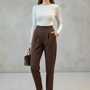 May include: A woman wearing a white long-sleeve shirt with black trim and brown high-waisted trousers. She is holding a small brown handbag and wearing nude-colored heels. The outfit is a classic, elegant style.