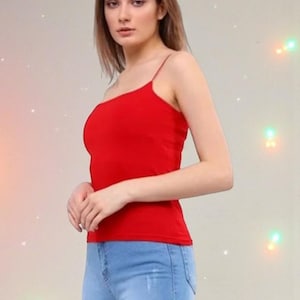 May include: A red camisole top with thin spaghetti straps. The top is paired with light blue denim jeans. The model is standing in front of a neutral background with soft lighting.