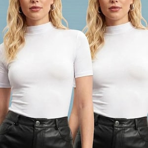 May include: Two women wearing white short-sleeve mock neck shirts and black leather pants. Both women have long blonde hair and are wearing silver hoop earrings. The background is a light blue color.