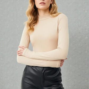 May include: A beige long-sleeved turtleneck top paired with black leather pants. The top has a close fit, and the pants have a sleek, polished appearance. The model is wearing gold hoop earrings.