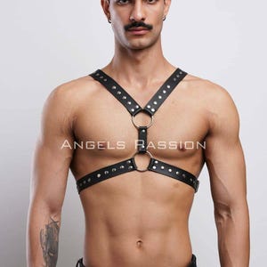 May include: Black leather harness with silver-tone rivets and metal rings. The harness has straps that cross over the chest and connect at the shoulders and waist. The text "ANGELS PASSION" is printed on the chest.