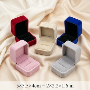 Könnte beinhalten: Fünf offene Ringboxen in verschiedenen Farben: Rot, Beige, Blau, Rosa und Grau. Jede Box hat ein schwarzes Innere und einen Schlitz für einen Ring. Die Boxen sind aus weichem, samtigem Material. Maße: 5 x 5,5 x 4 cm.