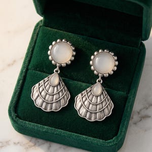 May include: A pair of silver-toned earrings featuring a round, milky white cabochon stone set in a scalloped frame, suspended above a textured, fan-shaped silver pendant with a small, teardrop-shaped white stone at the top center.