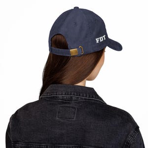 May include: Navy blue corduroy baseball cap with the embroidered letters "FDT" in white. The cap has a curved peak, a brass-coloured metal clasp, and an adjustable strap. The hat is a classic style.