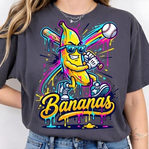 May include: Dark grey t-shirt with a cartoon banana wearing sunglasses and playing baseball. The banana is holding a bat and hitting a baseball. The word "Bananas" is written in a colourful, stylised font.