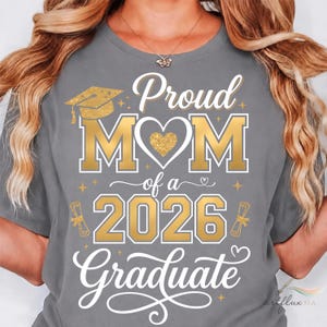 May include: A grey t-shirt with gold text reading "Proud MOM of a 2026 Graduate." The design features a graduation cap, heart, and diploma illustrations. The shirt has a crew neck.