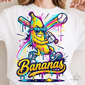 May include: A white t-shirt featuring a graphic of a yellow banana character wearing sunglasses, holding a baseball bat, and standing near a baseball. The design includes colourful paint splatters and the word Bananas in yellow script.