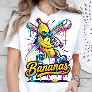 May include: White t-shirt with a cartoon banana wearing sunglasses and playing baseball. The banana is yellow with blue, pink, and green accents. The word "Bananas" is written in a yellow, dripping font.
