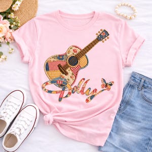 Può includere: Una t-shirt a maniche corte rosa chiaro con la grafica di una chitarra acustica con un motivo patchwork. La parola Dolly è scritta in un carattere corsivo con un design patchwork abbinato, accompagnata da una piccola grafica a farfalla.