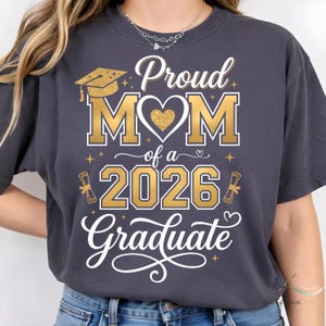 May include: Dark grey t-shirt with gold text reading "Proud MOM of a 2026 Graduate." The design features a graduation cap, heart, and scroll. The shirt is a casual style.