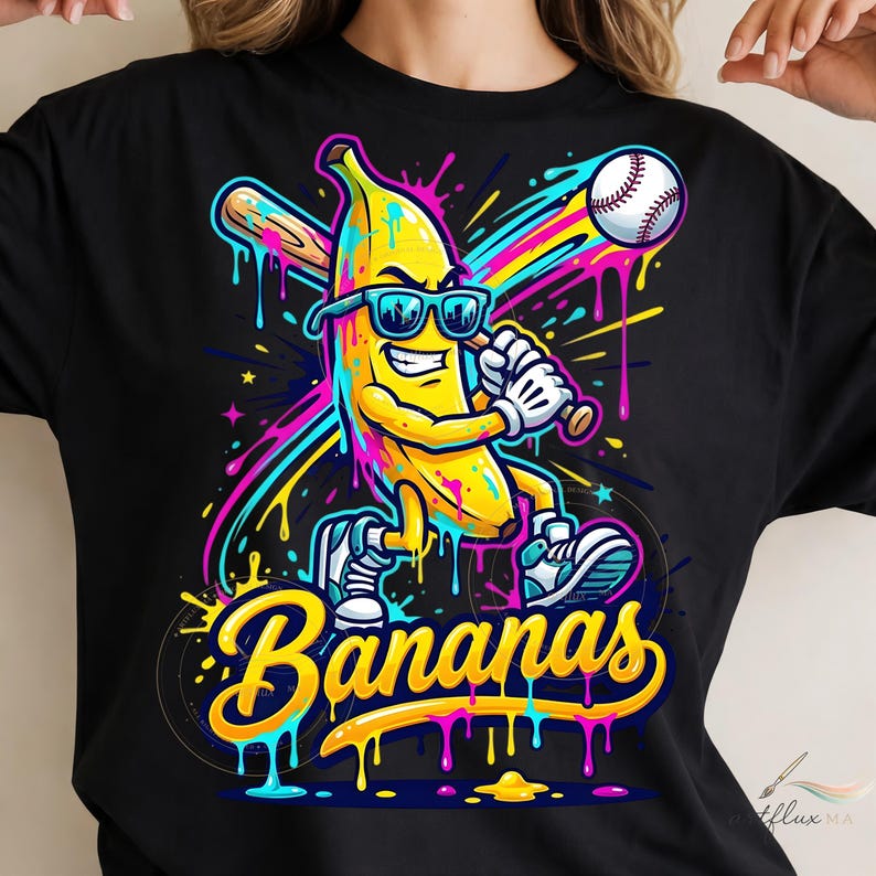 May include: A black t-shirt featuring a vibrant graphic of a yellow banana character wearing sunglasses, holding a baseball bat, and standing near a baseball. The design includes colourful paint splatters and the word Bananas in yellow script.