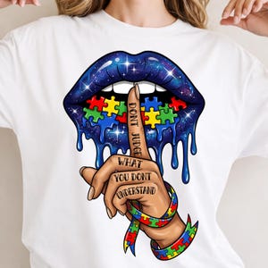 May include: White t-shirt featuring a graphic design. The design includes a pair of blue lips with puzzle pieces, a finger to the lips with the words "DON'T JUDGE", and a puzzle piece wristband. The words "WHAT YOU DON'T UNDERSTAND" are also visible.