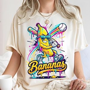 Può includere: T-shirt color crema con una banana dei cartoni animati con occhiali da sole che gioca a baseball. La banana è gialla con scarpe blu e una mazza da baseball. La parola "Bananas" è scritta in un font colorato e gocciolante.