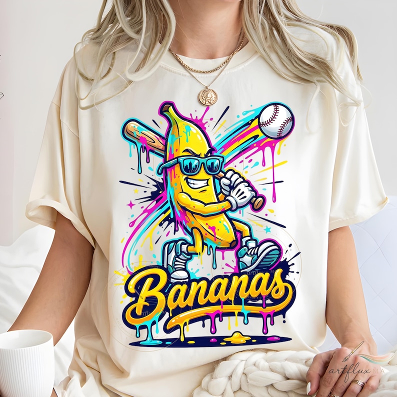 May include: Cream-coloured t-shirt featuring a cartoon banana wearing sunglasses and playing baseball. The banana is yellow with blue shoes and a baseball bat. The word "Bananas" is written in a colourful, dripping font.