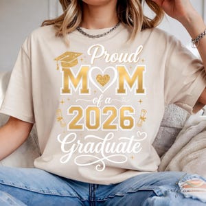 May include: Beige t-shirt with a gold glitter design that reads "Proud Mom of a 2026 Graduate." The design includes a graduation cap, heart, and scroll. The shirt is made of a soft material and is suitable for everyday wear.