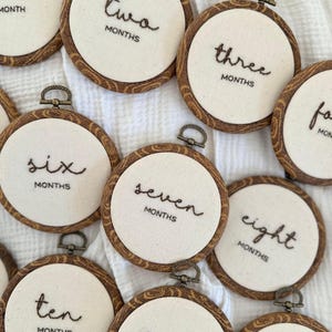 May include: Embroidered hoops with months of the year in black thread on cream fabric. The hoops have a wood grain design and metal clasps. The text includes 'one', 'two', 'three', 'six', 'seven', 'eight', 'ten', 'eleven'.