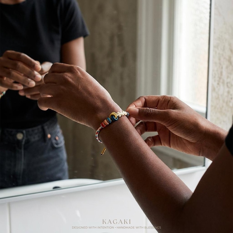 May include: A colorful beaded bracelet with a gold-colored clasp is being adjusted on a wrist. The bracelet features a rainbow of colors, including red, yellow, blue, and white. The word "KAGAKI" is visible at the bottom of the image.