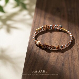 May include: A handcrafted bracelet with a woven design in shades of brown, white, orange, and blue. The bracelet features a central knot and a clasp. The word "KAGAKI" is printed at the bottom of the image, along with the text "DESIGNED WITH INTENTION &bull; HANDMADE WITH INTENTION."